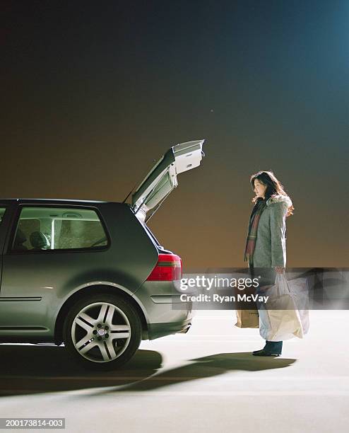 young woman holding bags, standing by open trunk of car, at night - trunk fotografías e imágenes de stock