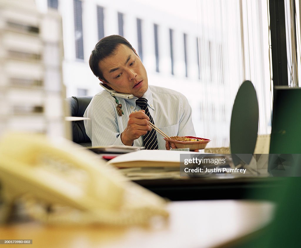 Businessman eating lunch at desk, using mobile phone