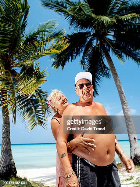 mature couple on beach smiling, woman embracing man from behind - fat guy on beach fotografías e imágenes de stock