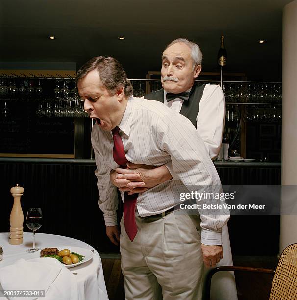 waiter with arms around choking man's stomach in restaurant - atemhilfe stock-fotos und bilder