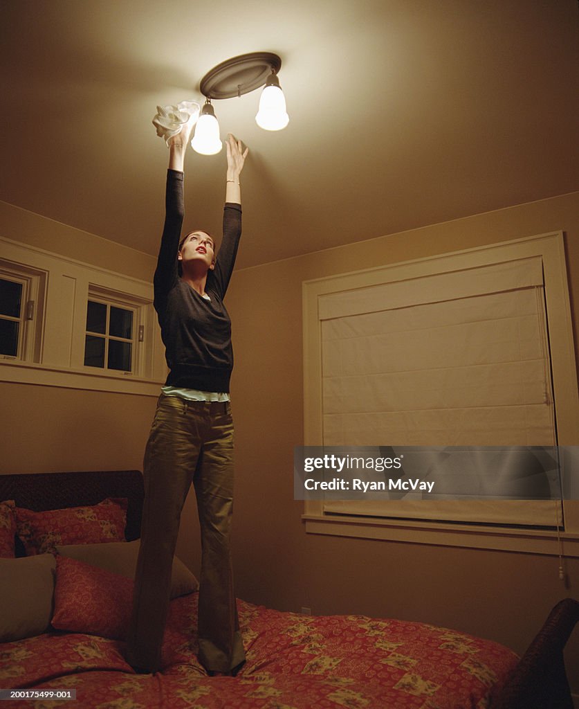 Young woman standing on bed to dust light fixture