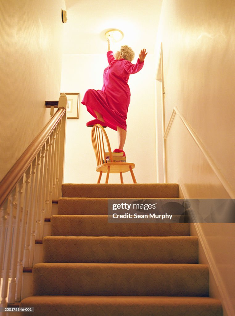 Senior woman changing light bulb above staircase, balancing on chair