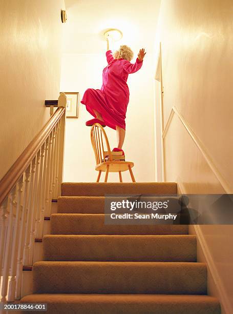 senior woman changing light bulb above staircase, balancing on chair - 不注意 個照片及圖片檔