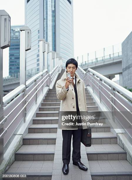 young businessman standing on steps, holding mobile phone - オーバーコート ストックフォトと画像