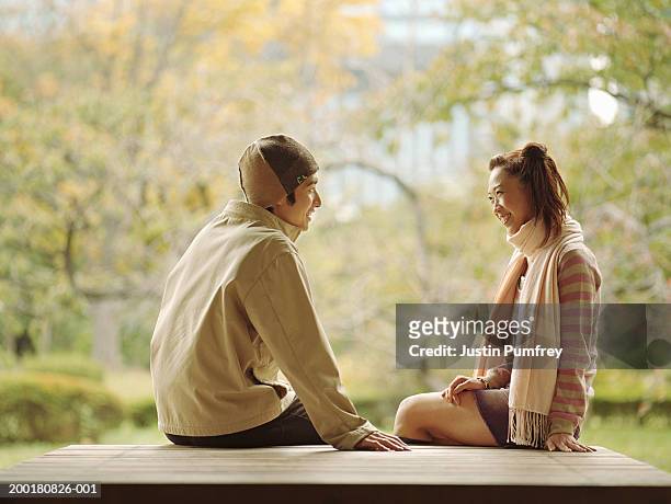 young couple sitting outdoors, facing each other, smiling, side view - asian man seated stockfoto's en -beelden