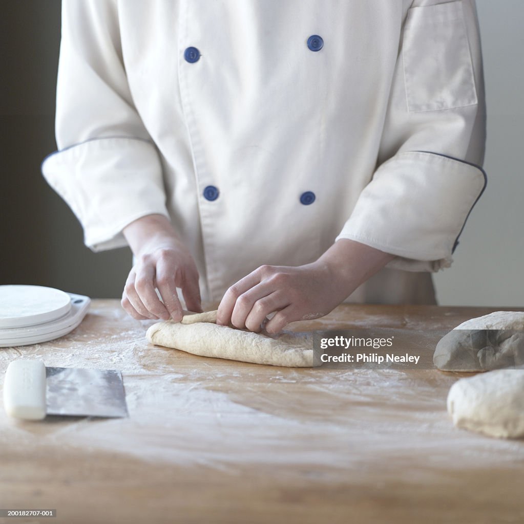 Young woman baker folding dough on floured work surface, mid section