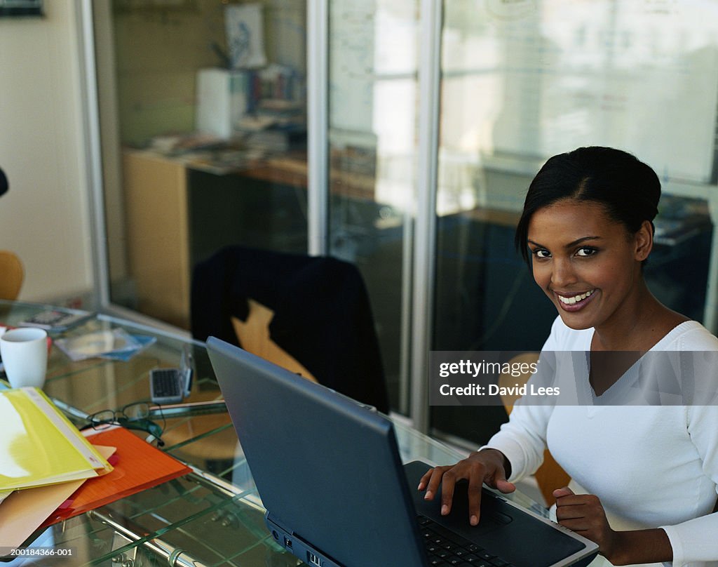 Young woman using laptop in office, smiling, portrait