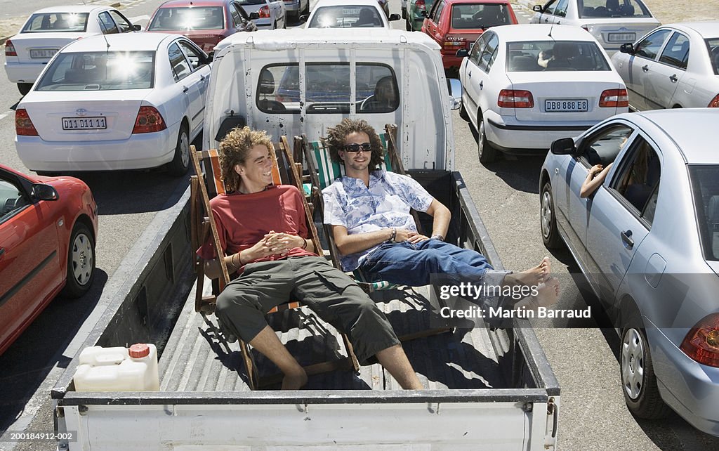 Two men on deckchairs in back of pickup truck amongst traffic jam