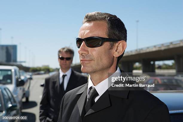 two mature businessmen, wearing sunglasses outdoors by cars - lijfwacht stockfoto's en -beelden