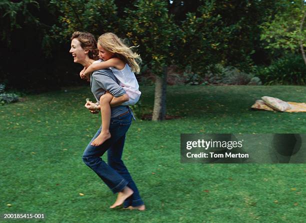 woman giving daughter (5-7) piggyback, running on lawn - portare a cavalluccio foto e immagini stock