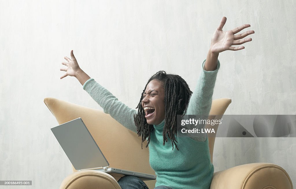 Young woman, sitting in armchair with laptop, arms outstretched