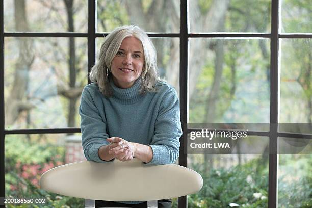 mature woman leaning of back of chair indoors by window, portrait - une seule femme d'âge mûr photos et images de collection