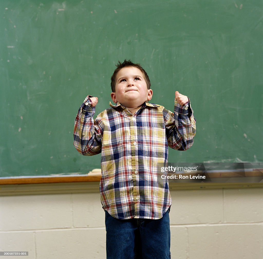 Boy (3-5) with fists closed standing in front of blackboard