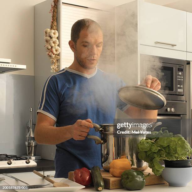 man in kitchen lifting lid on saucepan emitting steam - deksel stockfoto's en -beelden