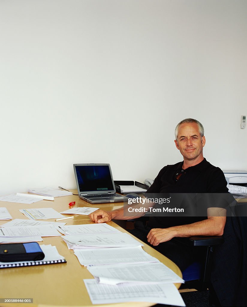 Mature man sitting at desk covered with papers, smiling, portrait