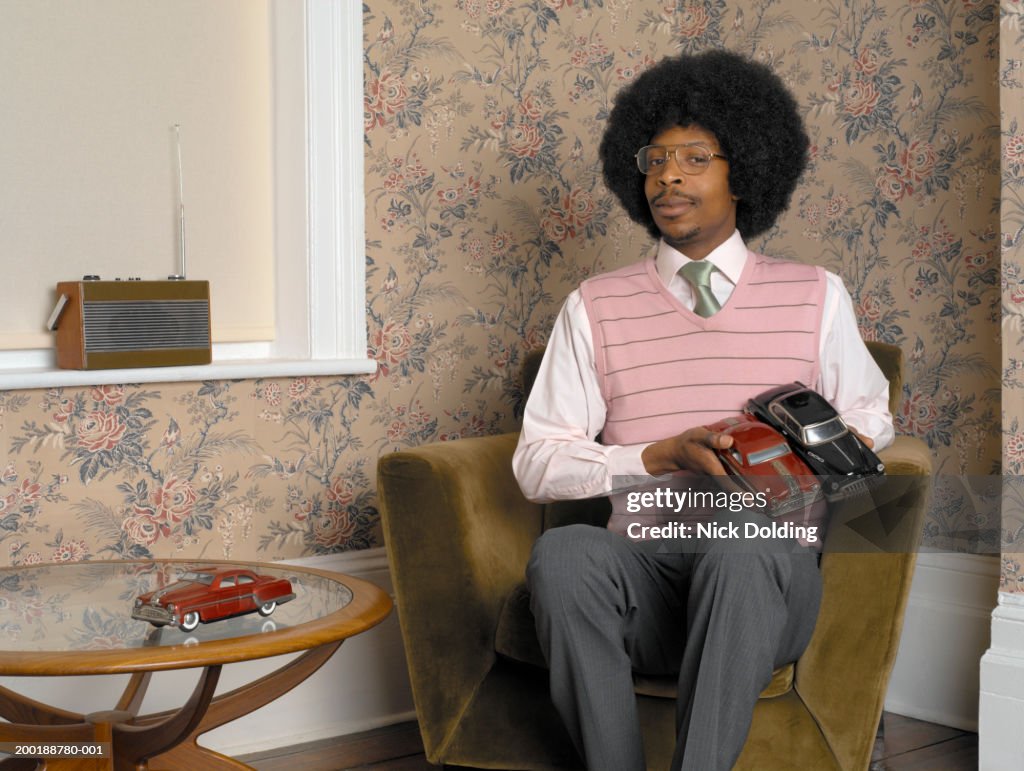 Young man sitting in living room, holding model cars, portrait
