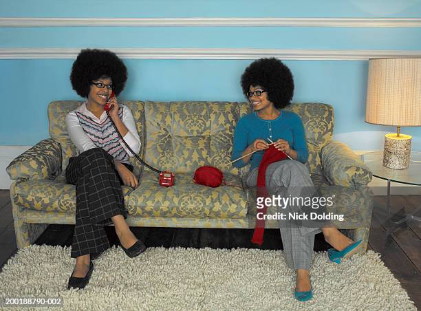 two young women on sofa, one using telephone, other knitting - grey trousers stock-fotos und bilder