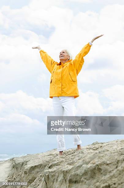 senior woman on rock wearing slicker at beach, raising arms upwards - yellow trousers stock-fotos und bilder