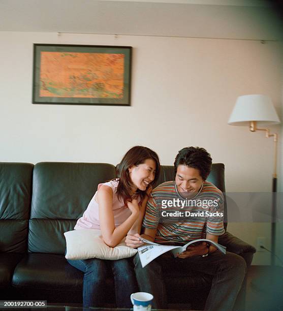 young couple on couch smiling, man looking at magazine - picture magazine stockfoto's en -beelden