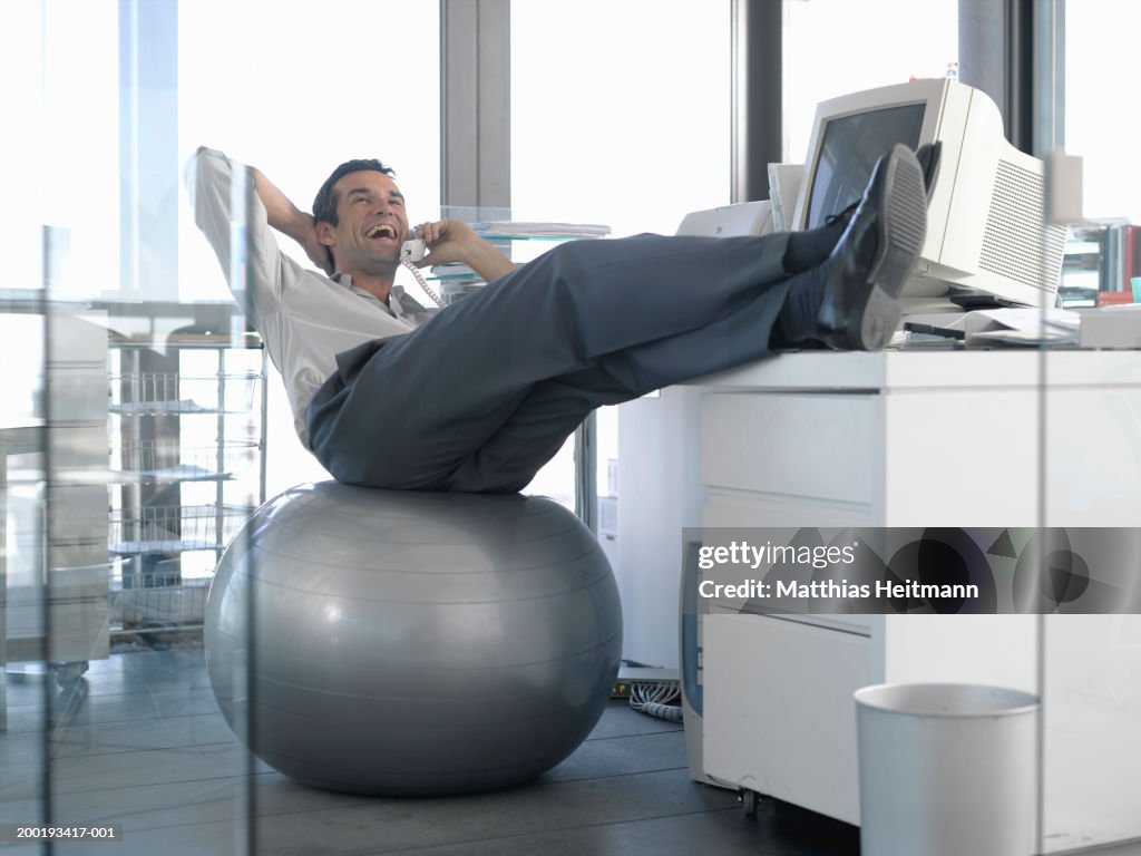 Businessman sitting on excercise ball at desk, using phone, laughing