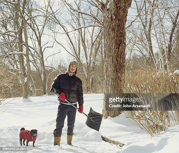 man standing in snow with dog, holding shovel, portrait - snow shovel man stock pictures, royalty-free photos & images