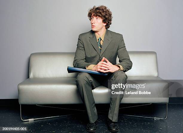 young man sitting on sofa, looking off to side - vorstellungsgespräch stock-fotos und bilder