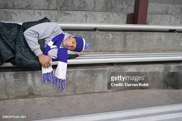 young football fan asleep in sleeping bag on stadium bench - soccer scarf stock-fotos und bilder
