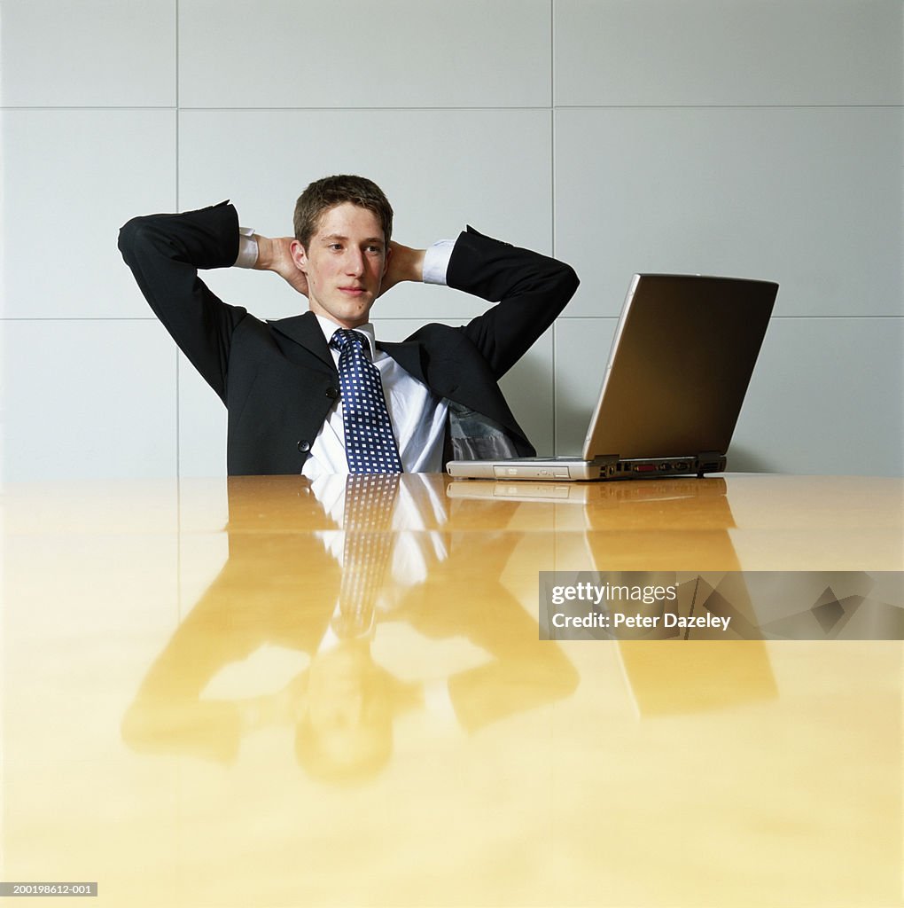 Teenage boy (17-19) wearing suit, hands behind head, looking at laptop