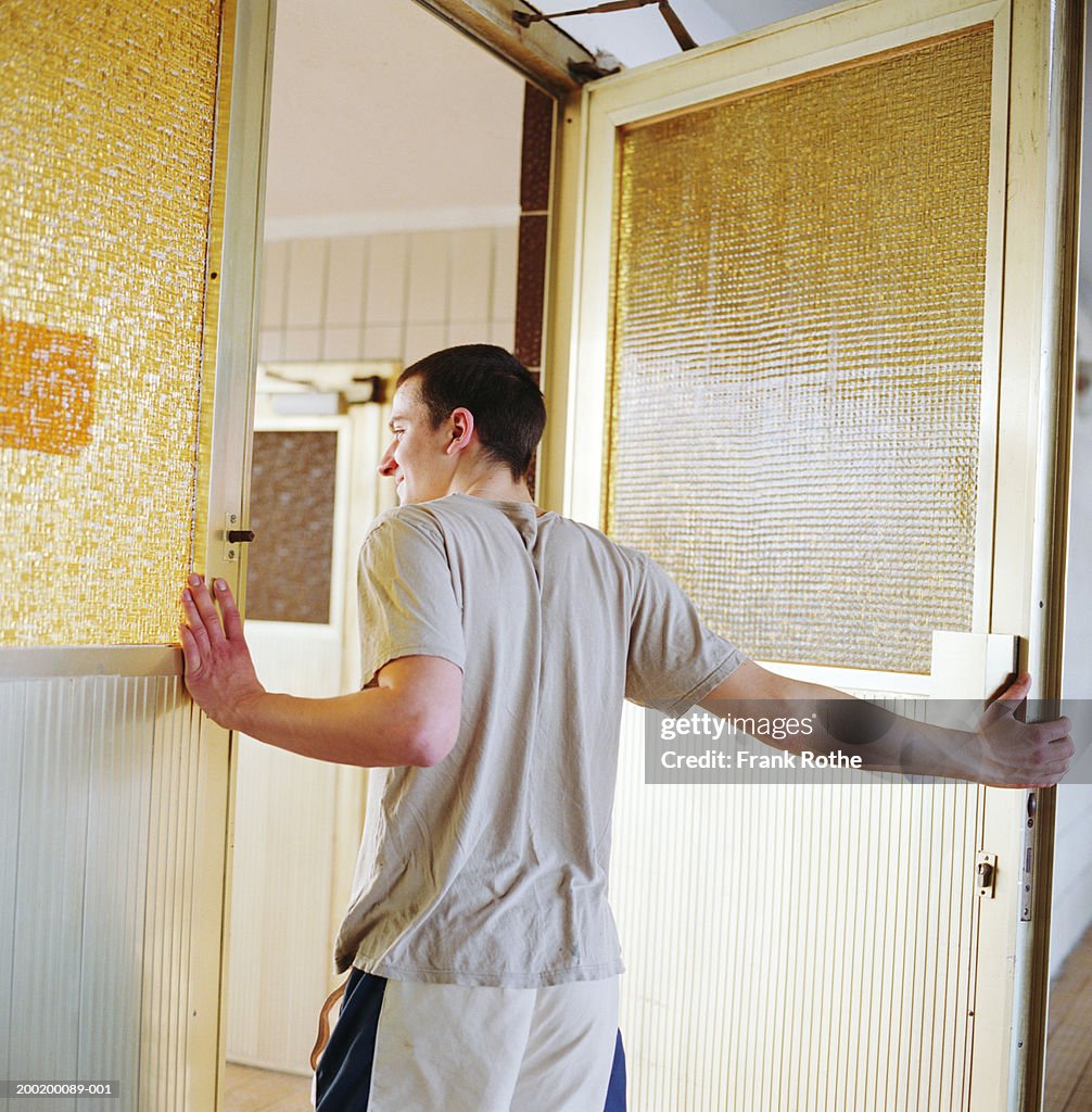 Young man holding door open, peering out, rear view