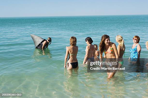 man wearing mock shark fin on back by group of people in sea, laughing - plastikhai stock-fotos und bilder