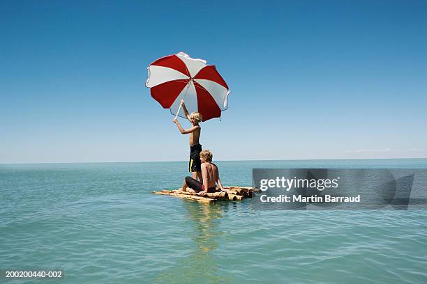 two boys (10-12) on bamboo raft in sea, one holding up parasol - floß stock-fotos und bilder