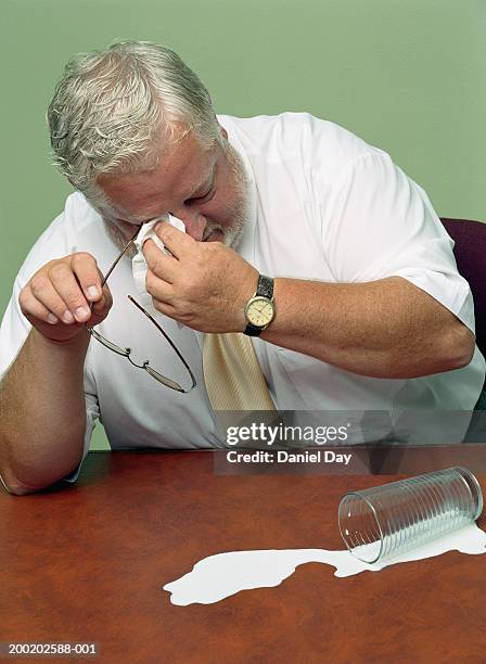 man sitting at desk with glass of spilt milk, wiping eyes - spilt milk foto e immagini stock