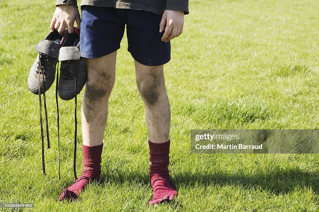 Boy (8-10) standing in field, holding football boots, low section