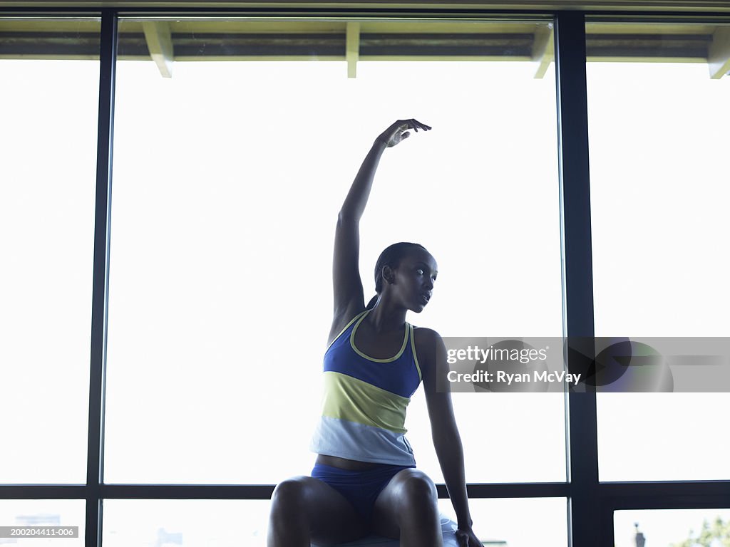 Young woman sitting on fitness ball, arm raised, looking away