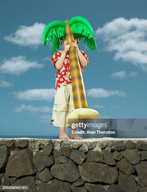 boy (3-5) standing on wall holding inflatable palm tree - aufblasbarer gegenstand stock-fotos und bilder