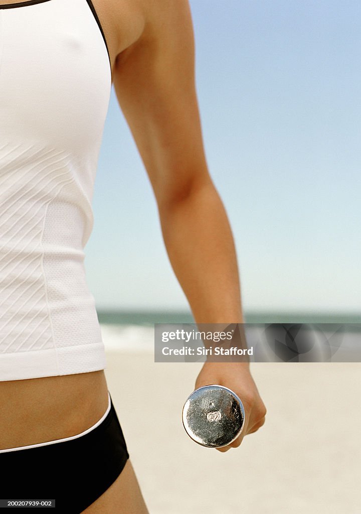 Young woman holding dumbbell on beach (mid section)