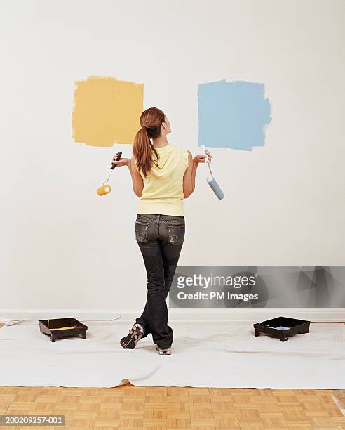 young woman holding paint rollers, looking at wall, rear view - wand streichen stock-fotos und bilder