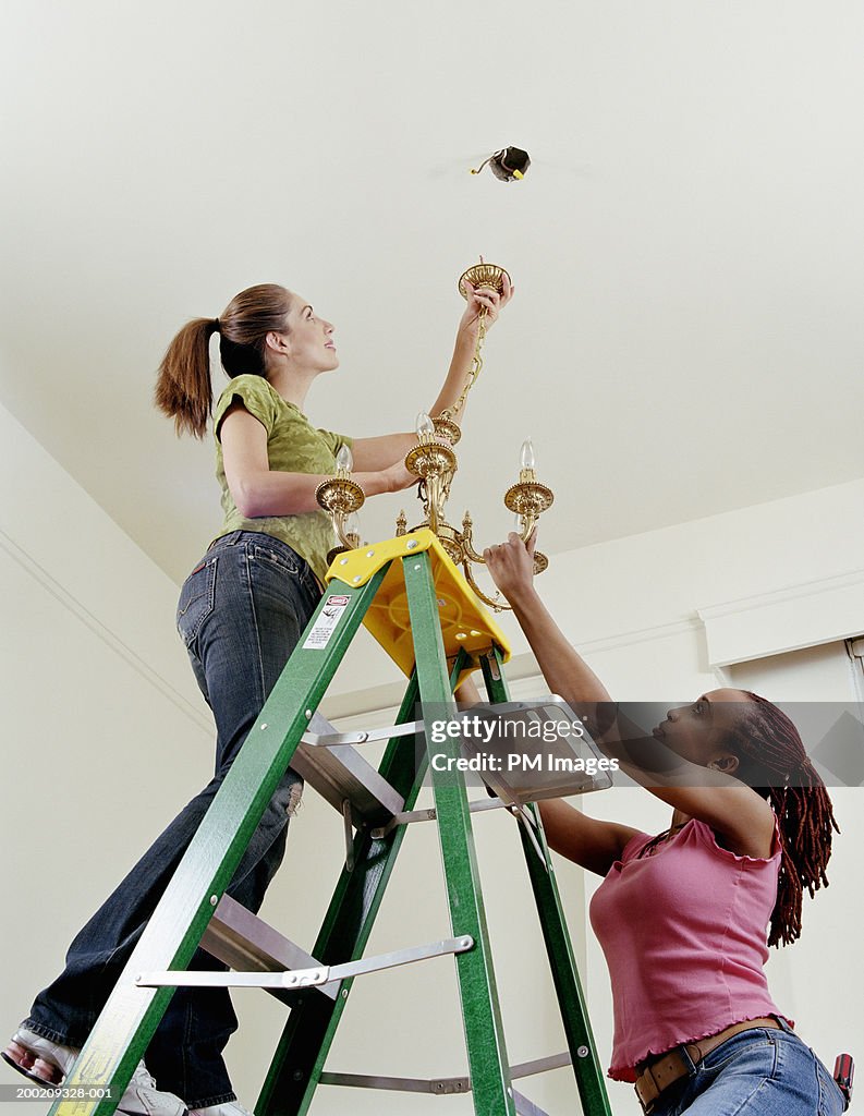 Two women on ladder hanging light fixture to ceiling