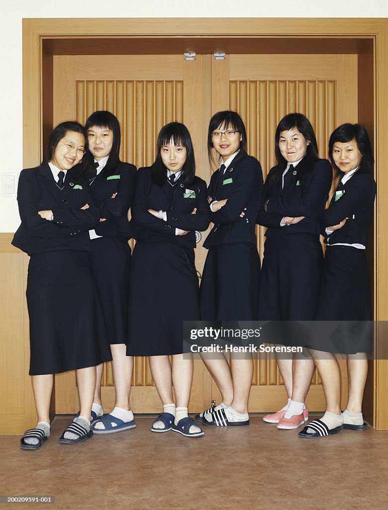 Six schoolgirls (14-16) standing by doorway, arms folded, smiling
