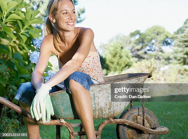 young woman sitting in wheelbarrow, looking away, laughing - wheelbarrow stock pictures, royalty-free photos & images