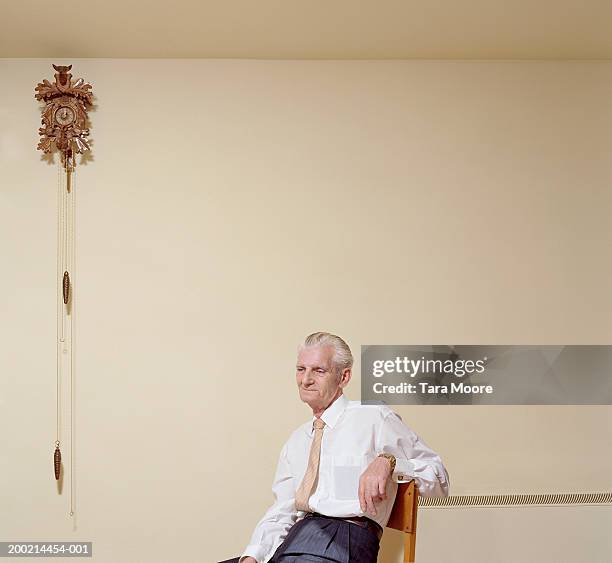 senior man in shirt and tie, sitting on chair by cuckoo clock - cuckoo clock stock pictures, royalty-free photos & images