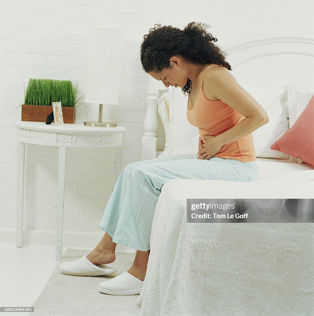 Woman sitting on bed holding stomach, head bowed