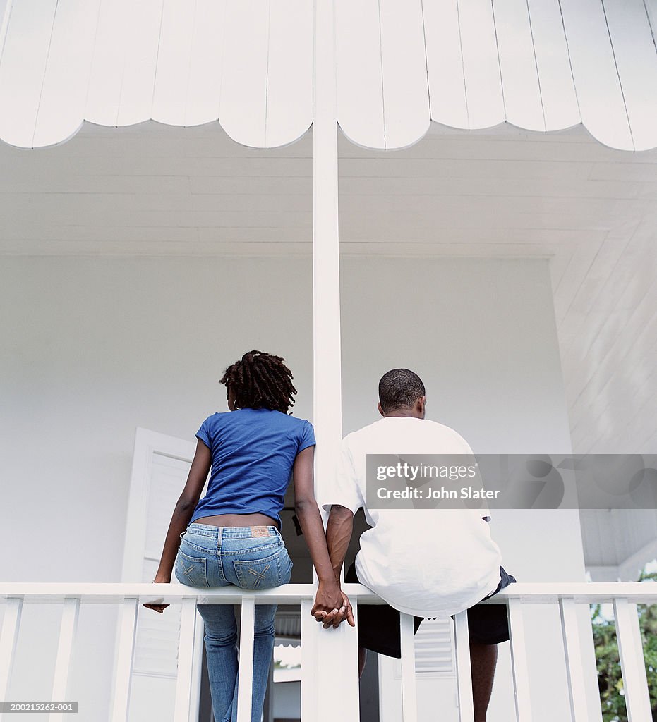 Couple relaxing on veranda, sitting on rail, holding hands, rear view