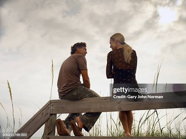 couple sitting on ledge near beach at sunset, side view - beach fence stock-fotos und bilder