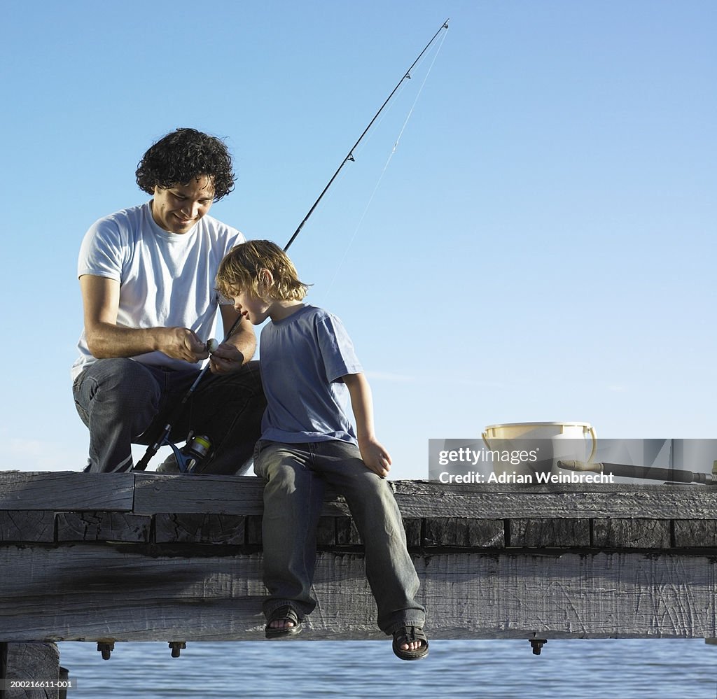Father showing son (4-6) how to bait fishing hook on jetty, smiling