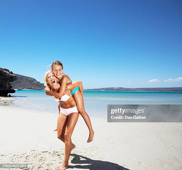 mother giving daughter (8-10) piggyback on beach, laughing - fille maillot photos et images de collection