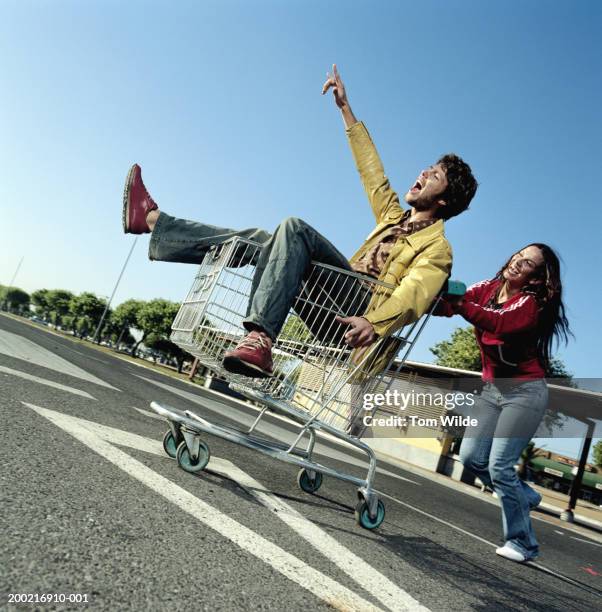 young woman pushing young man in shopping trolley in carpark - two point stock-fotos und bilder