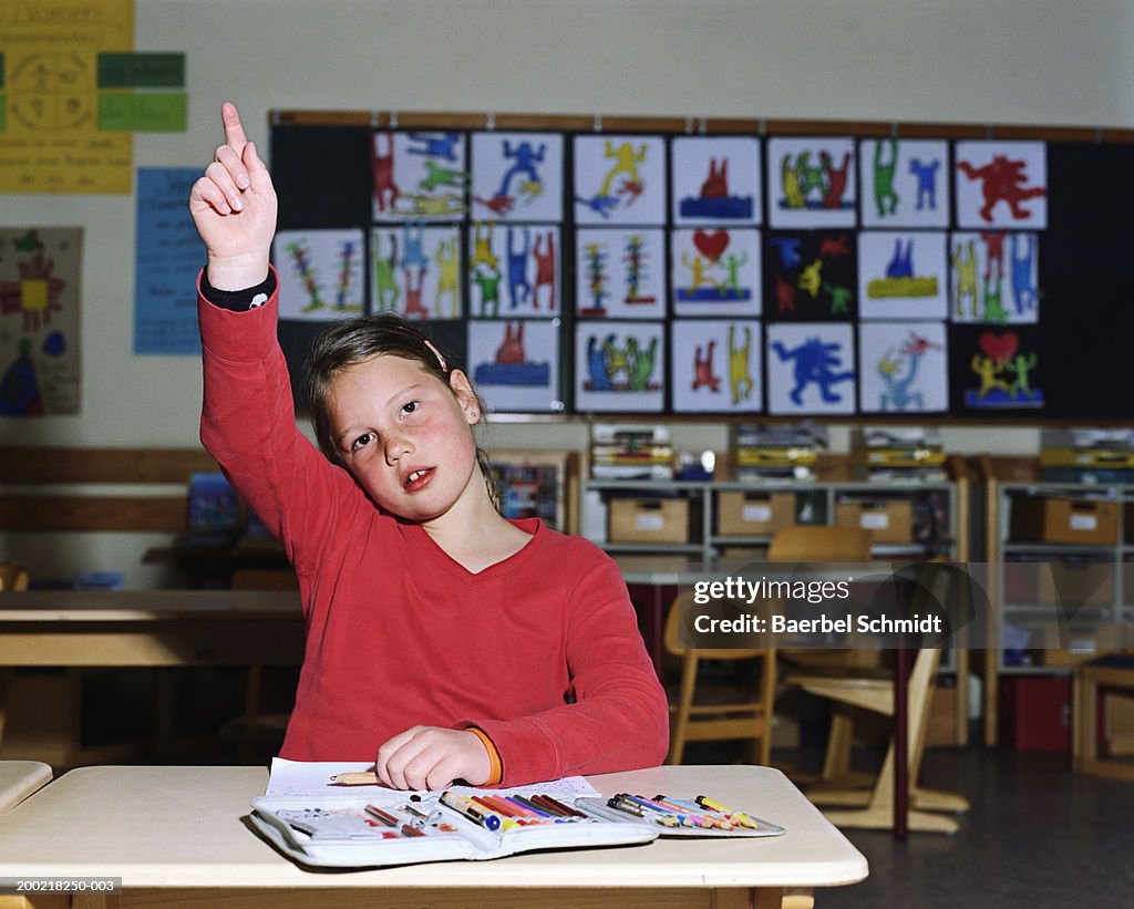 Girl (8-10) at desk raising hand in classroom, portrait