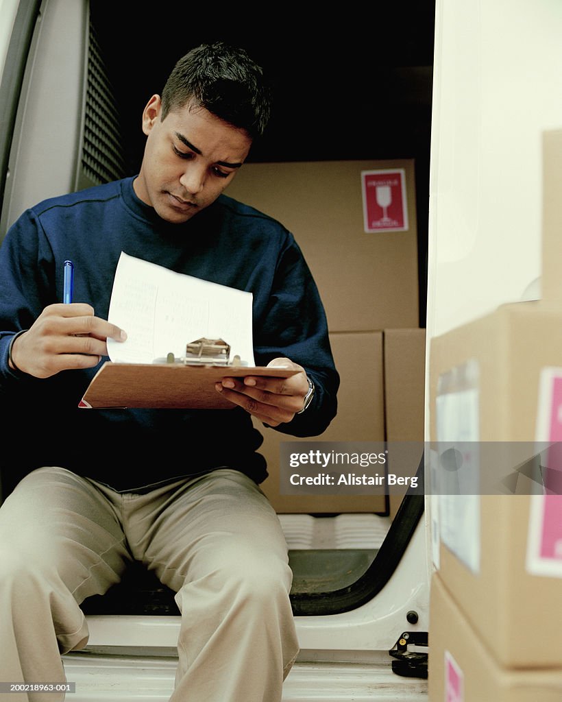 Young delivery man sitting on back of van looking at clipboard