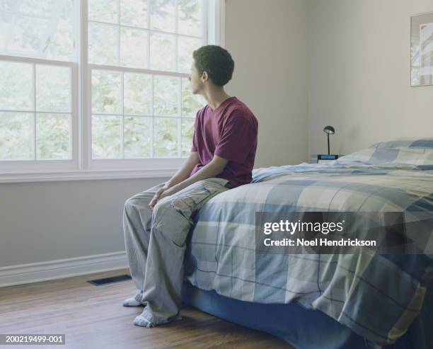 teenage boy (16-18) sitting on edge of bed, looking out window - boy sitting on bed stock-fotos und bilder